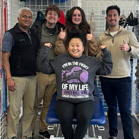 Patient Kaitlynne sitting on therapy table at RWW outpatient facility while flexing and smiling. Her four therapists are standing behind her smiling and giving thumbs up.