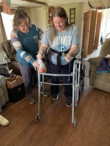 Patient Theresa in her living room standing behind her walker while also holding a bag of oranges. Her therapist in standing behind her on the right side.