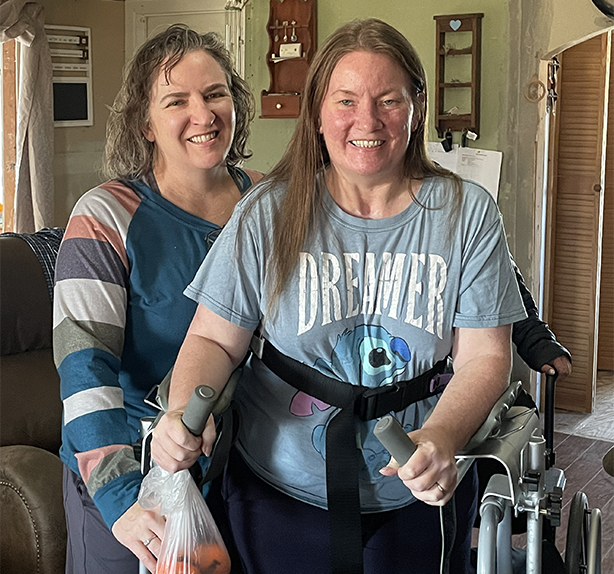 Patient Theresa in her living room standing behind her walker while also holding a bag of oranges. Her therapist in standing behind her on the right side.
