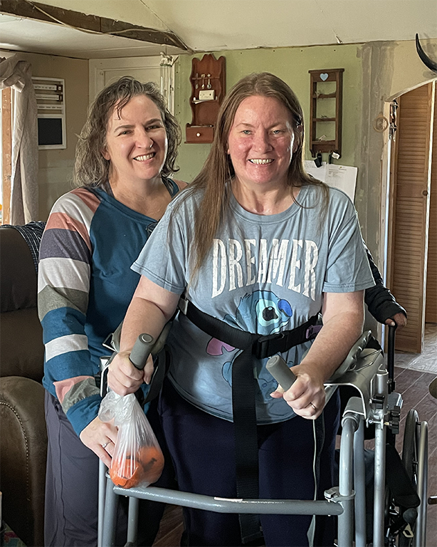 Patient Theresa in her living room standing behind her walker while also holding a bag of oranges. Her therapist in standing behind her on the right side.