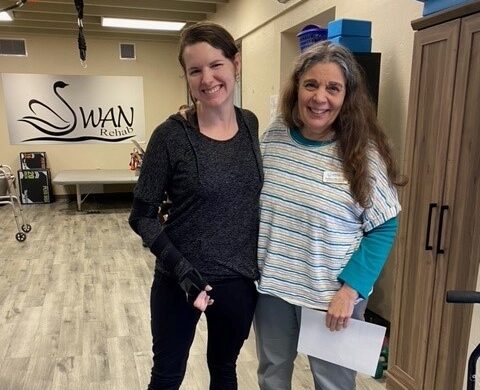 Patient Rebekah standing in outpatient center with device on her arm. Standing next to her therapist.