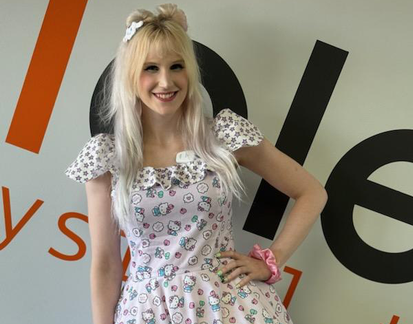 Patient Ariana standing in front of a wall with hand on hip wearing a light pink dress smiling. she has blonde hair.