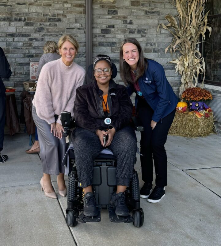 Two white women standing behind young african american woman sitting in power wheel chair. All smiling.