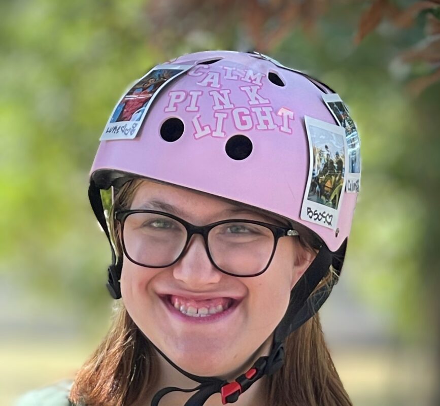 Young white woman with long brown hair smiling wearing glasses and a pink helmet with the saying calm pink light on it.