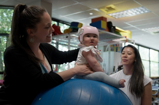 Woman therapist is smiling while holding baby on a blue therapy ball. Baby is wearing pink head gear. Another therapist is on the other side of the ball looking at the baby.