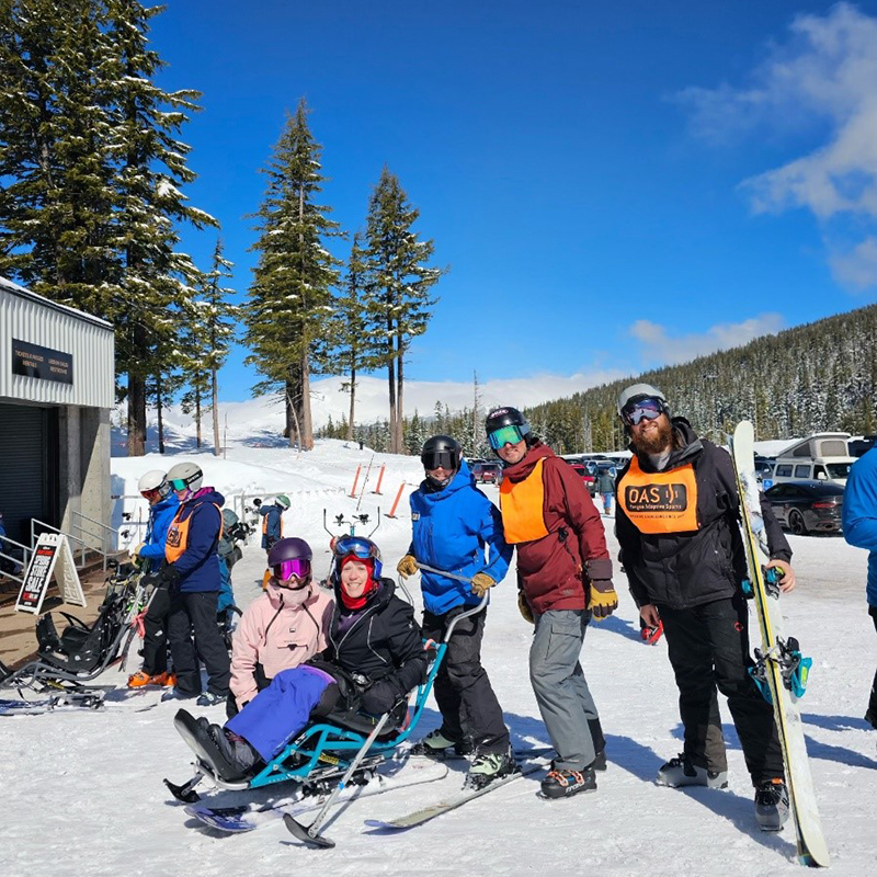 Patient Barbara sitting in adaptive skiing seat wearing helmet and full ski outfit. Standing behind her are three therapists on skis wearing helmets and reflective ski goggles. There is a sunny blue sky and snow covered trees and ground.