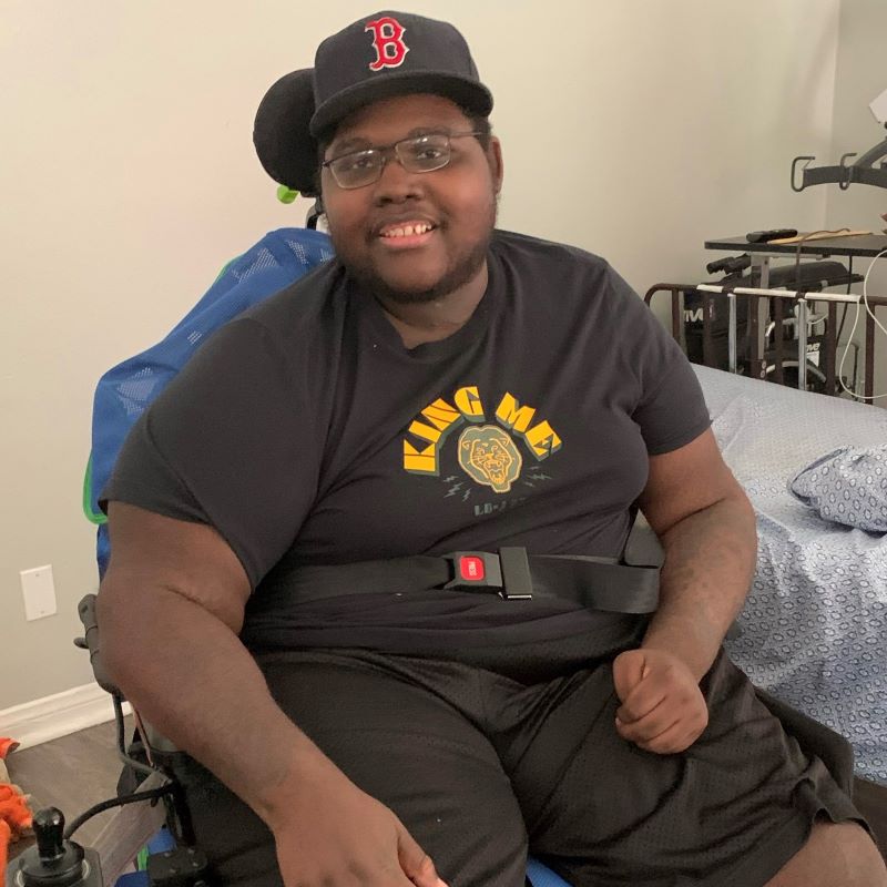 Mr. Goins is wearing glasses and a black baseball cap, with a black tshirt and shorts. he is sitting in a power wheelchair.