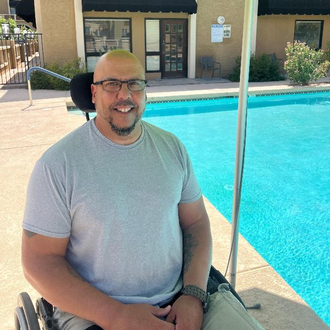 Mr. Ramirez is sitting in his wheelchair smiling and is next to a pool with blue water.