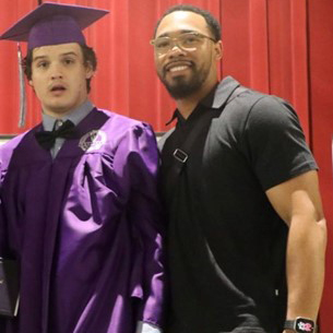 Colby is wearing a purple hat and gown over a black tuxedo. He is standing on the left of his caretaker who is wearing glasses, a black shirt, and smiling.