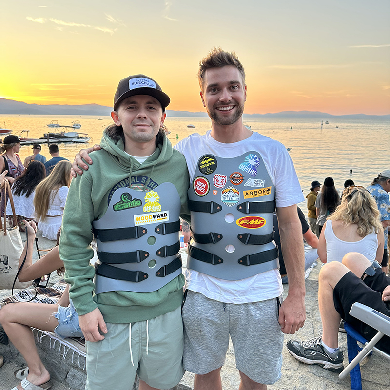 Patient Alvaro standing next to his friend with lake in the background and a sunset mixed with yellow and orange. Person on the right is wearing grey shorts and white t-shirt with brace around his torso. Person on the left is wearing a black hat with green sweatshirt and grey shorts with a brace around his torso as well. Both men are smiling.