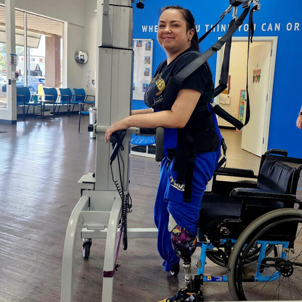 Patient Bree is standing in front of a wheelchair in a harness wrapped around her upper body by a machine. she has both legs amputated and one prosthetic leg. she is smiling.