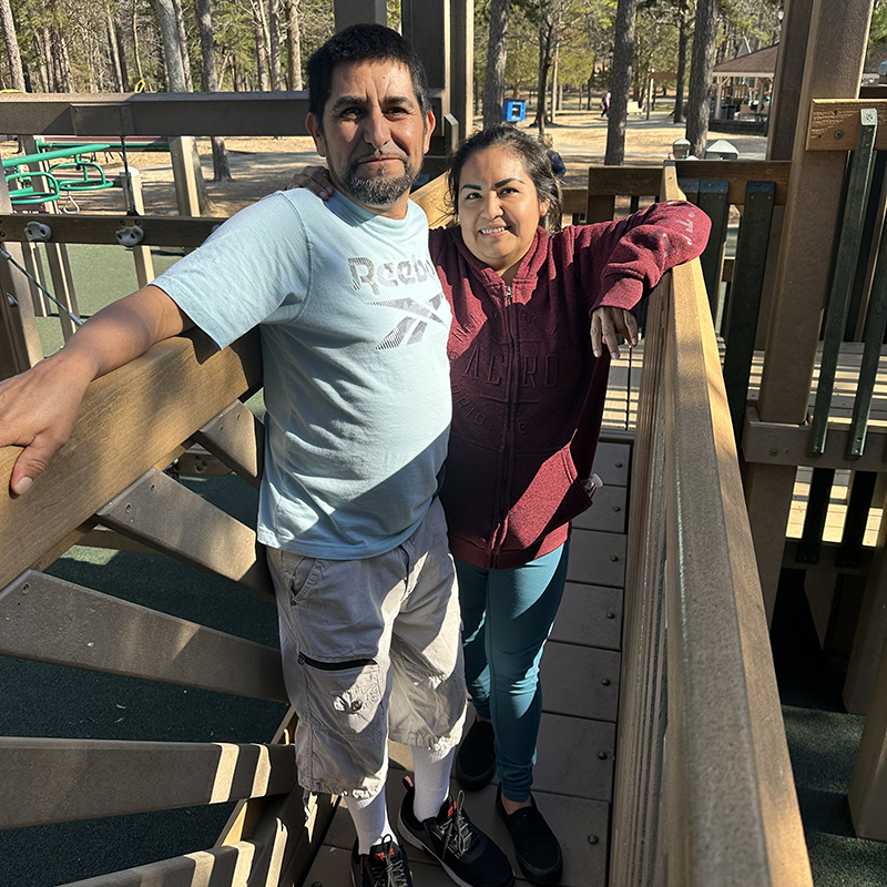 Patient Jorge with his wife standing in the park on a bridge. Both smiling.