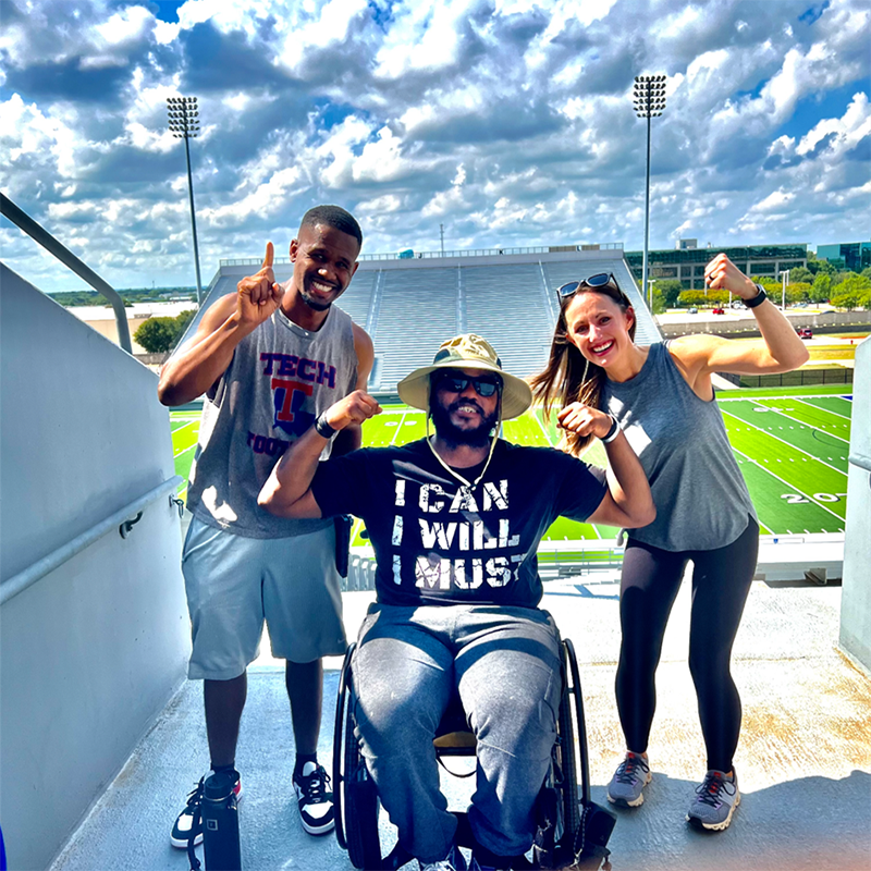 Patient Michael is sitting in his wheelchair wearing a large bucket hat, with his arms up making fists and flexing. Two people are standing on each side of him also flexing and smiling. they are all standing in a stadium with the field behind them.
