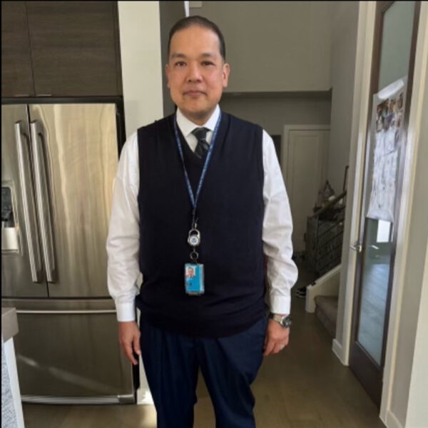 Patient Juan is standing in his kitchen with his hands by his side smiling. He is wearing navy slacks and a black vest and tie with white shirt underneath.