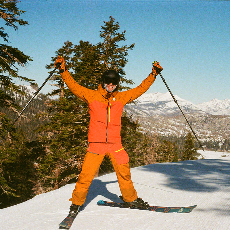 Alex is in an orange ski outfit with a helmet and goggles. He is on skis and holding ski poles in the air and smiling. He is on top of a mountain with trees and more snow covered mountains in the background.