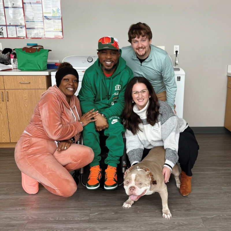 Brandon is wearing a green jumpsuit with orange sneakers sitting down in his wheelchair. His wife is next to him kneeling down wearing a velvet pink jumpsuit. The rehab director is behind him and Community Relations Manager is kneeling besides him holding Brandon's bulldog.