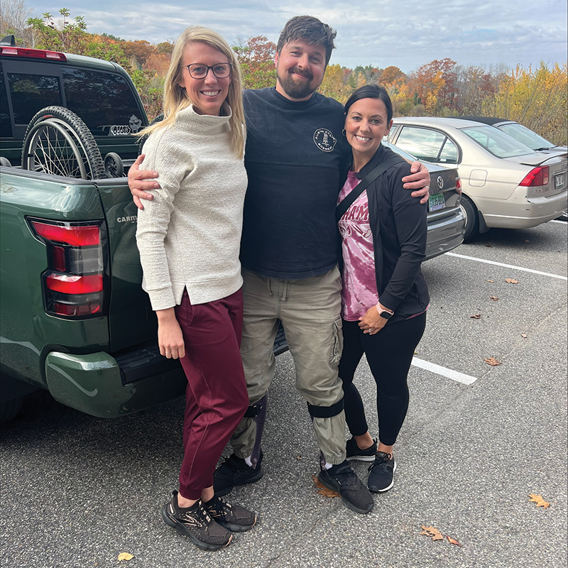 Patient Sean is standing in the middle of his two therapists who are both women. He is wearing ankle-foot orthosis on both feet.