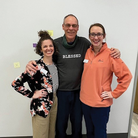 Patient Tim is standing in the middle of his two therapists. He is smiling, wears glasses, and wearing a dark green sweatshirt with the words blessed on it. The therapist to the right of him is a woman wearing an orange sweatshirt and glasses. The therapist to his left is a woman with dark curly hair smiling. She is wearing a black cardigan with bright flowers.