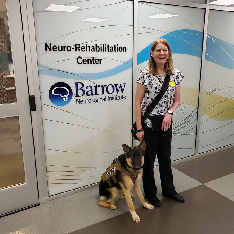 Patient Lori is standing with her German Shepard dog sitting next to her. Lori is smiling, she has light blonde hair.