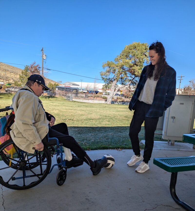 Patient Craig G. is sitting in his wheelchair while kicking around a soccer ball with his therapist.