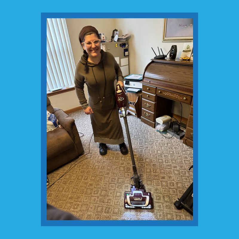 Client Kim B. is standing in her house smiling while holding a vacuum cleaner.