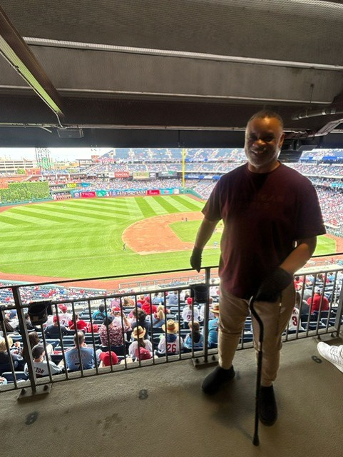 Kenneth S. is standing with his cane, smiling, in front of the Phillies baseball diamond inside the stadium.