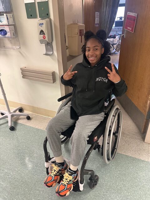 Mh’Chiya is an a young African American girl, sitting in a wheelchair, smiling and making a peace sign with her hands.