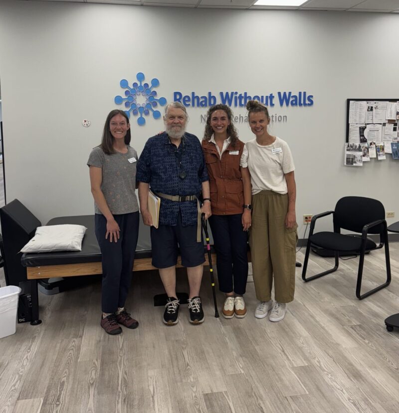 Phil is an older gentleman with a grey beard holding a cane, standing with three of his female therapists at his side.