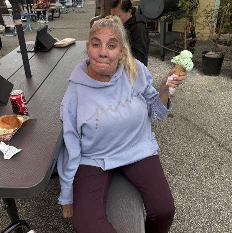 Patient Heather is sitting down at a table outside with an ice cream cone in her hand. She has blonde hair and is wearing a purple sweatshirt and dark purple pants.