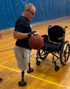 Crash is a middle-aged white male, double leg amputee wearing leg prosthetics. He is standing dribbling a basketball on a basketball court. He is wearing glasses, a black t-shirt, and grey shorts.