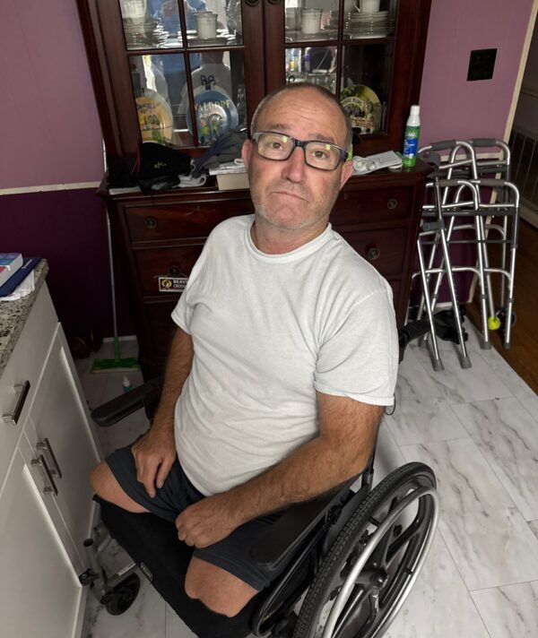 Crash is a middle-aged white male, double leg amputee sitting in his wheelchair wearing a white t-shirt and smiling at the camera. He is wearing glasses.