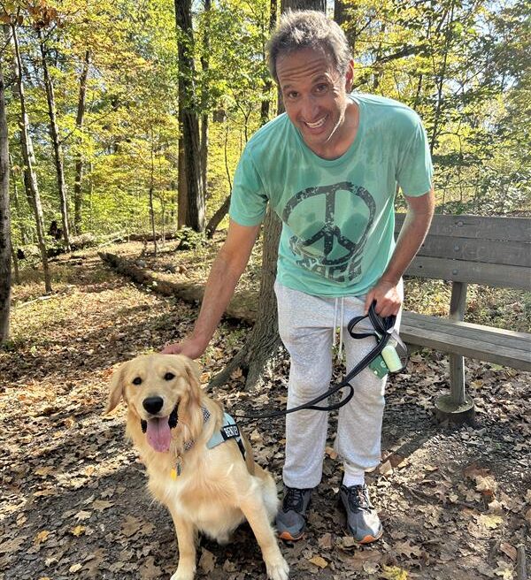 Mike and his golden retriever dog are standing on a trail with a lot of trees and leaves around them.