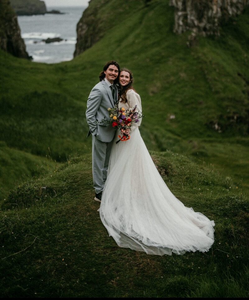 Max and his wife are standing on gassy hills with the sea and large rocks behind them. They are dresses in traditional wedding attire with a suit and white wedding dress.