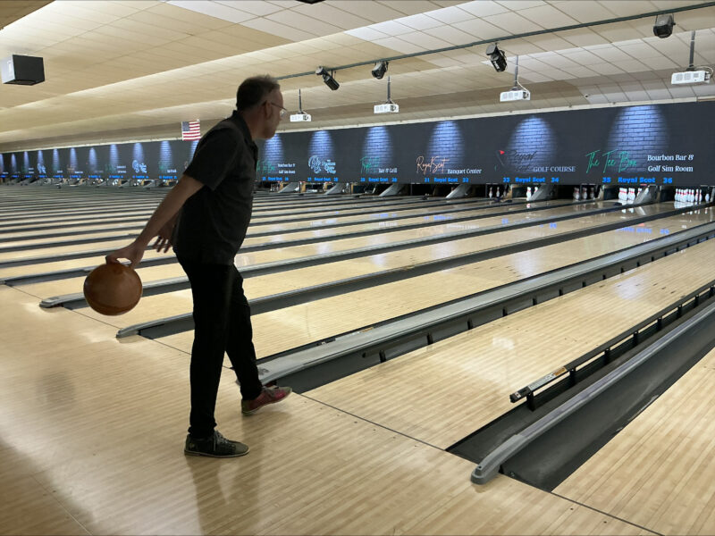 Greg is at a bowling alley, standing right bewfore the lane start with bowling ball in his right hand about to roll the ball down the lane.