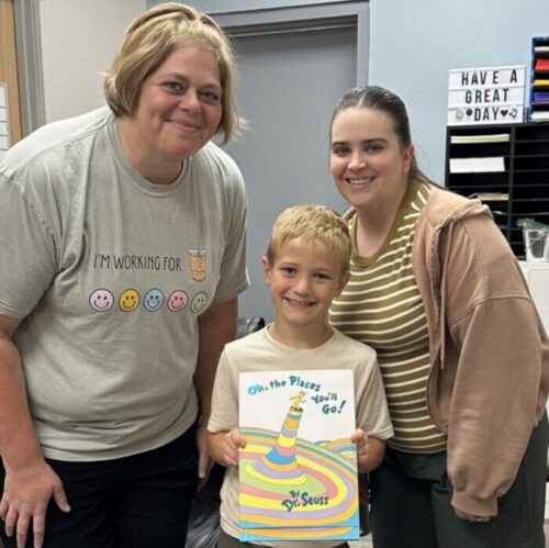 Two adult therapists smiling behind a child also smiling holding a Dr. Suess book.