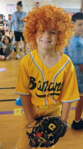 Small child wearing an orange curly wig, a yellow jersey, and holding a baseball glove.